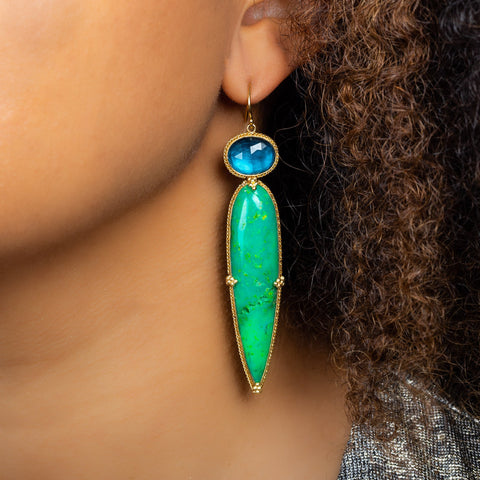 A model wears a large earring featuring an oval shaped blue topaz atop a teardrop shaped green turquoise stone. 