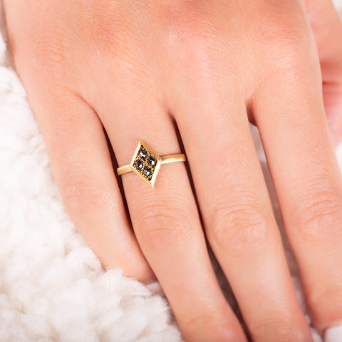 A model wears a diamond shaped ring that is crafted with four small chain wrapped black diamonds and set in an 18k yellow gold bezel. The ring is set on a thin band.