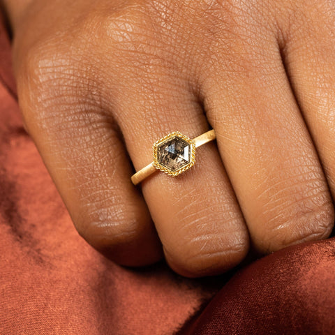 A model wears a small hexagon shaped charcoal colored diamond in an 18k yellow gold setting on a thin ring band.