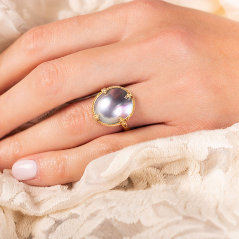 A model wears a large iridescent natural pearl ring with a chain wrapped 18k yellow gold bezel and four beaded prongs on a thin band