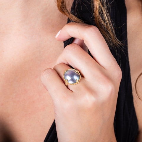 A model wears a large pearl ring with grey, blue and purple hues in a chain wrapped bezel with four beaded prongs on a thin band