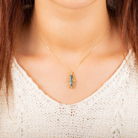 A model wears an irregular oval shaped opal and wood pendant is set in an 18k yellow gold chain wrapped bezel with four beaded prongs. The pendant hangs on a short delicate chain.