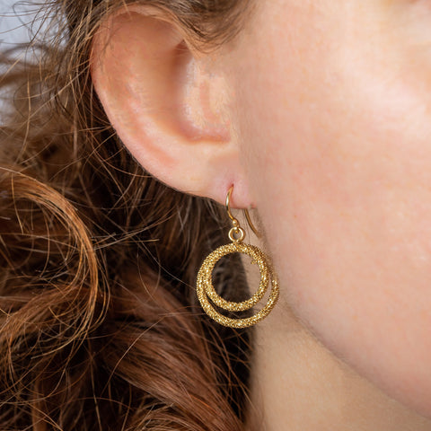 A model wears a small 18k yellow gold earring featuring two interlocking circles crafted with chain to create a stardust-like effect.