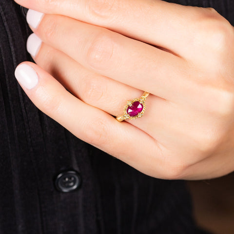 A model wears a small oval shaped ruby ring with a braided bezel and four granulated prongs on a thin 18k yellow gold band.
