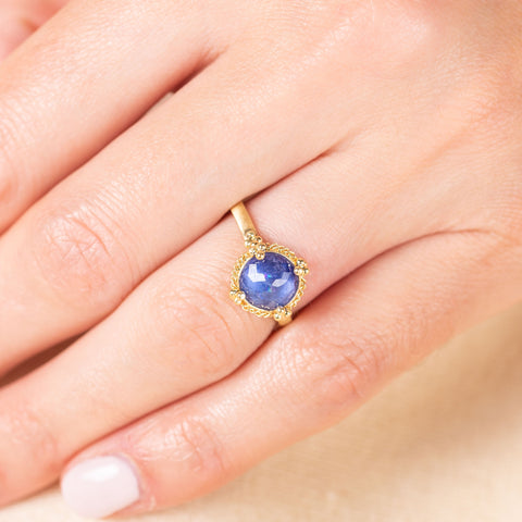 A model wears a faceted round tanzanite stone ring set in 18k yellow gold with a braided detail around the bezel and four granulated prongs.