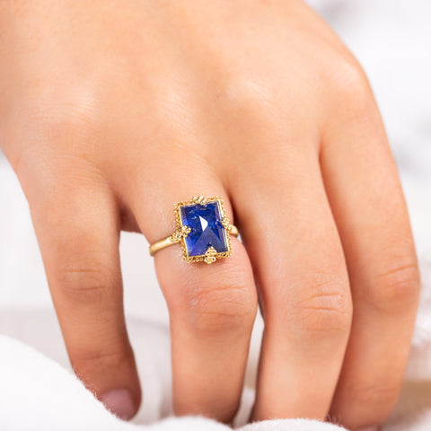 A rectangular deep blue Tanzanite ring is set in an 18k yellow gold chain wrapped bezel with four beaded prongs. The stone is set on a thin band.