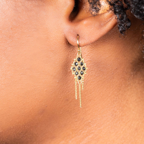 A model wears an 18k yellow gold earring crafted with black diamonds woven into a diamond shaped lattice pattern and has three dangling chains at the bottom.