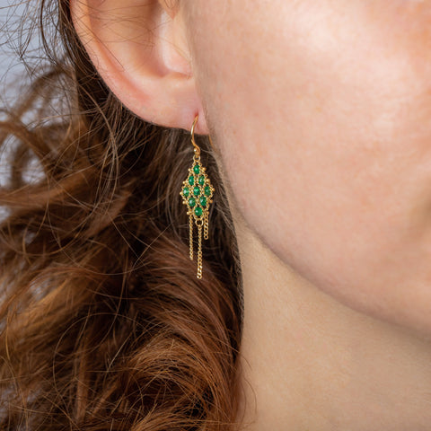 A model wears an 18k yellow gold earring crafted with green emeralds woven into a diamond shaped lattice pattern and has three dangling chains at the bottom.