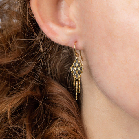 A model wears an 18k yellow gold earring crafted with grey diamonds woven into a diamond shaped lattice pattern and has three dangling chains at the bottom.
