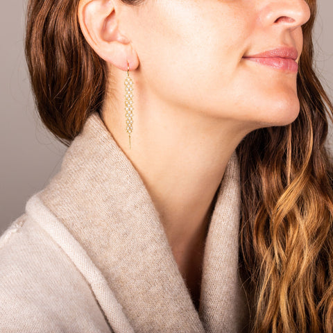 A model wears a long white pearl earring featuring a woven lattice pattern and two dangling chains at the bottom. The earring. fasten with french hook closure.